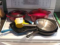 Full view of all cookware items arranged on stove: red enamelled cast iron covered pots, blue enamelled grill pan, stainless steel frypan, black non-stick frypan, and small yellow sauce pot.