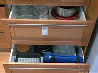 Two opened drawers showing clear glass casserole and mixing bowls, metal colanders, and a blue carrying case with burger patty press plus various metal bake tins inside the lower drawer.