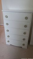 Full front view of the white mid century dresser showing five drawers with round knobs and a scalloped bottom drawer edge.