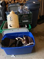 Wheeled Rubbermaid garbage container, several plastic totes with lids, green Heritage Farm crate with bags, beige water jug with spigot