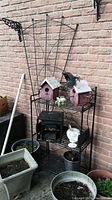 Full view of metal garden trellis behind a folding metal shelf with birdhouses, planters, and frog figurine on an outdoor patio against a brick wall.