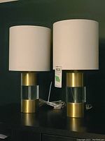 Pair of identical table lamps on dark surface showing full lamps with white drum shades and cylindrical brass and acrylic bases
