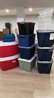 Stack of large Rubbermaid Roughneck style plastic totes in dark blue, gray, and black, two red and white Igloo coolers, and a blue plastic tool caddy.