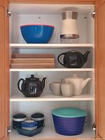Kitchen cupboard shelves showing teapots, cups, bowls, salad spinner, and kitchen organizers