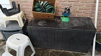 Black plastic yard storage bench with three stepstools stacked on and around it and a box with two green garden hoses on top.