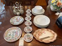 Photo of assorted Asian dinnerware including decorative plates, white plates and bowls, floral teacups with saucers, scalloped floral tray, and silver condiment containers on wooden table.