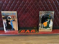 Three bar-themed decorative mirrors aligned on floor against padded leather wall. Central mirror smaller with BAR text, flanked by two larger artistic mirrors.