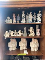 Full view of three shelves with multiple ceramic figurines including traditional dressed figures, cherubs, busts, and decorative vases.