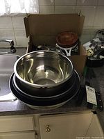 Photo showing nested stainless steel mixing bowls and black baking pans stacked on a kitchen counter next to sink.