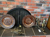 Copper plate hanging trays, two large round copper plates with embossed Victorian style scenes, set of horse medallion ornaments, two knight figurines, other metal ornamentals on black tiled hearth in front of brick fireplace.