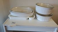 Six white CorningWare casserole bowls and three lids on a white surface. Bowls are stacked and arranged with some oval and some round shapes. Lids are clear glass, one with chip on edge.