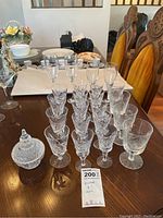 Side angle photo showing 16 stemmed wine glasses and 4 larger goblet glasses arranged in rows on dining table, with covered dish to the side.