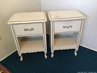 Two white wooden end tables with curved legs and scalloped edges, each with one drawer and an open shelf below, placed side by side against a wall.