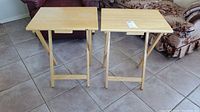 Two wooden folding tables with natural wood finish, visible signs of wear, positioned side by side on tiled floor next to upholstered furniture.