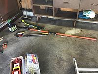 Overview of garden tools and equipment including large cutting tool with orange handle, moss control container, and boxed items on garage floor.