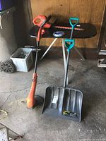 Photo showing full lot including snow shovel, weed eater, hedge trimmer, and trim line on garage floor and table.