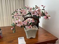 Glass bonsai tree with pink flowers displayed on a wooden surface, showing fine details of flowers and branches.