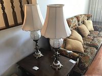 Pair of brass and glass lamps with fabric shades on a wooden table next to a sofa with floral upholstery and pillows.