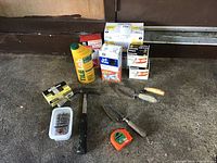 Photo showing all items including light bulbs, tile grout bottle, measuring tape, hammer, and three trowels arranged on the ground.