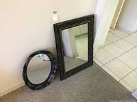 Photo showing two framed mirrors, one large rectangular and one smaller oval, both with ornate dark carved frames, leaning against a white wall.