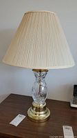 Full view of lamp showing cream pleated shade, clear glass body, brass tone base and lamp tag on table.