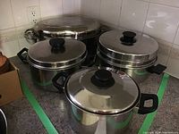 Photo showing three stainless steel pots with lids and black handles alongside a large turkey roaster with lid on a kitchen counter.