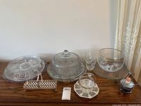 Overview of the lot including clear glass serving platter with dome, punch bowl and glasses, small clear glass bowl on doily, cobalt blue condiment jar with metal holder, and small woven basket.