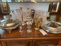 Wide shot showing all silver plated serving ware and decorative items including candelabra, goblets, sugar shaker, brass bucket, and serving dishes.