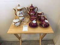 Two Japanese tea sets displayed on a small wooden folding table, one with floral gold-accented china, the other burgundy with gold trim, all pieces visible.