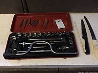 Open red case showing socket wrench set with ratchet, T-handle, extensions, and various size sockets along with tap and die pieces in top compartment.