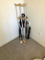 Two crutches paired with various umbrellas in a corner against a beige wall and carpeted floor.