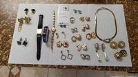Overview of jewelry lot including earrings, watches, rings, necklace, and cuff links laid on white paper background.