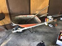 Full view of metal wheelbarrow with orange tray and light gray frame, sitting indoors against wood panel wall.