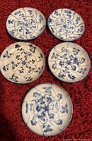 Top-down view of five antique Chinese blue and white porcelain saucers arranged on a red textured surface showing detailed floral and vine hand-painted patterns.