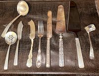 Full set of nine assorted sterling silver flatware pieces laid flat on a brown textured surface, showing both plain and ornate handles and different types of serving utensils.
