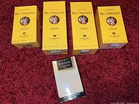 Group of five unopened fragrance boxes on red textured surface: four yellow Bal A Versailles and one Shalimar Guerlain box.