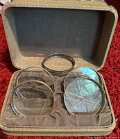 Set of four round glass coasters with sterling silver rims, arranged in original gray cushioned box with zippered closure on red carpet.