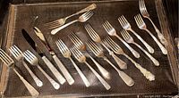 Full display of assorted forks and some knives arranged on a brown textured surface showing various handle designs and tarnish.