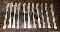 Twelve plain handled sterling silver butter knives arranged in one row on textured brown background