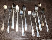 Twelve assorted sterling silver forks laid out on a textured dark mat, showing varying handle styles and fork tines.