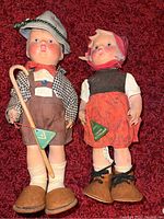 Photo of two vintage Hummel dolls laid side by side on a red carpet showing one doll in lederhosen with green hat and cane and the other in a red skirt with headscarf and brown shoes.