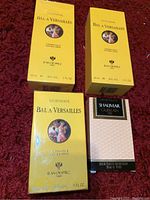 Four unopened perfume packages placed on a red surface, showing three yellow Jean Desprez Bal A Versailles and one pink Guerlain Shalimar box.