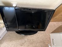 Top-front angled view of the 32 inch black Dynex TV on carpet, showing screen, stand, and power cable draped over screen.