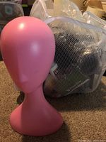 Pink mannequin head on carpeted floor with a clear mesh storage bin with hair and other items inside in background