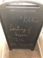 Front side of one black plastic-framed chalkboard A-frame with chalk writing advertising an event 'Eclectic Kitchen tasting & bazaar 2-6p, Food, Sangria, Flowers'