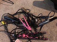Five plug-in hair styling tools including flat irons and a hot comb tangled with cords on carpet.