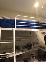Photo of white metal loft bed with blue mattress cover and integrated desk below. Ladder attached on left side. Room has various items beneath the desk.