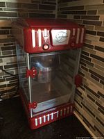 Front view of red popcorn maker with clear glass door, metal heating compartment inside, red door latches, and Crofcor branding on front panel.