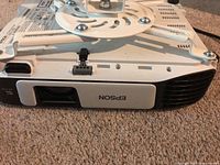 Front view of the Epson projector showing the lens, some vents, and part of the ceiling mount bracket attached on top.