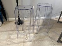 Two clear acrylic ghost-style bar stools side by side on tiled floor, showing transparent body and legs with visible scuffs and scratches.
